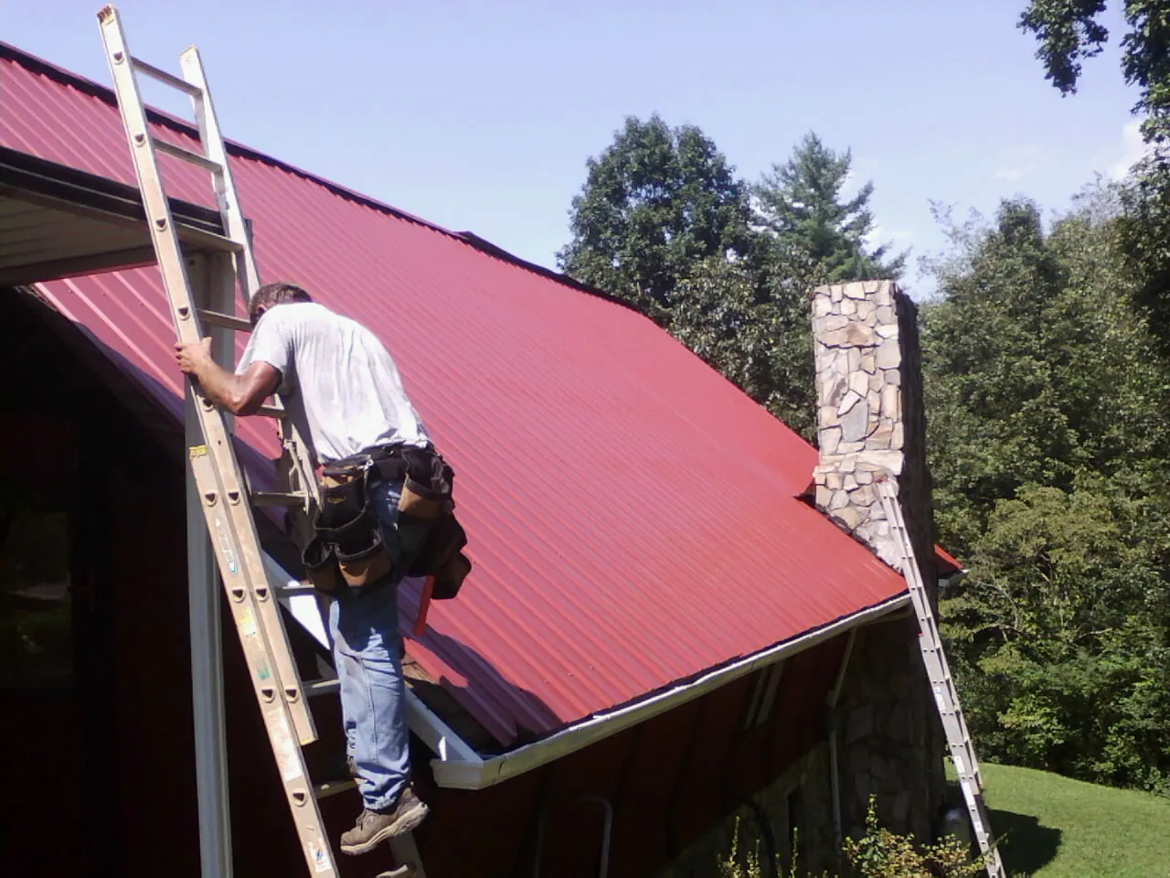 Roofer on a ladder beside a red metal roof and stone chimney — Soffit & Fascia Repair by Roofing Brothers in Tussy